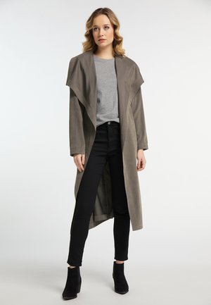 Femme aux cheveux blonds ondulés portant un long manteau taupe par-dessus une chemise grise, un pantalon noir et des bottines noires, debout sur un fond blanc.