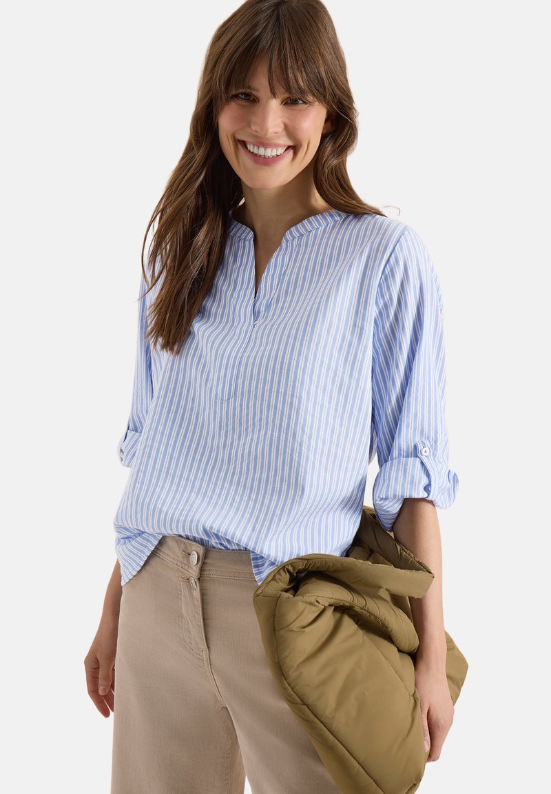 Femme souriante, portant un chemisier rayé bleu clair et blanc avec des manches retroussées, un pantalon beige, et tenant une veste vert olive pliée.