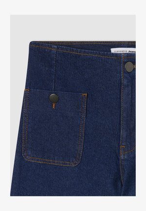 Jeans in denim blu scuro con una tasca frontale dotata di un bottone in metallo nero e cuciture gialle a contrasto lungo i bordi. Texture complessivamente ordinata.
