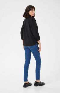 �Černá blůza s třičtvrtinovými rukávy a sklady, v kombinaci s modrými slim-fit džíny a černými mokasíny, na jednobarevném pozadí.