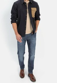 Marineblå skjorte med knaplukning og en beige brystlomme, beige T-shirt under, blå jeans og brune lædersko. Glat tekstur, afslappet pasform.