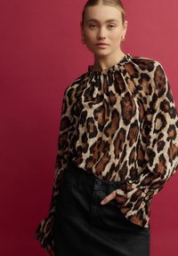 Halenka s leopardím potiskem, smršťovacím výstřihem, dlouhými rukávy a rozšířenými manžetami, spárována s černou koženou sukní. Pozadí v jednolité barvě.