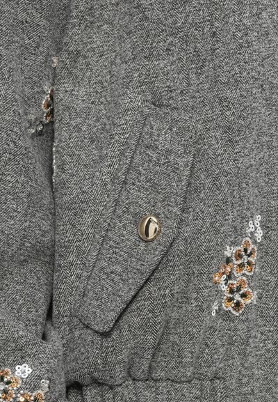 Pelēks zīda audums ar kabatu ar atloku, kuram ir zeltraņa poga un ziedu rakstu secinājumi sudrabkrāsā, melnā un bronzas krāsā.