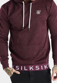 Uomo con felpa con cappuccio color bordeaux screziata con cordoncini bianchi e la scritta "SIKSILK" sull'orlo, con le mani parzialmente nelle tasche dei pantaloni neri.