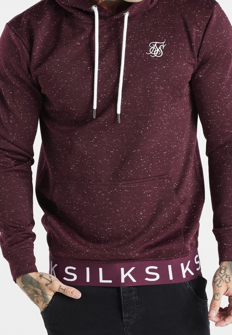 Uomo con felpa con cappuccio color bordeaux screziata con cordoncini bianchi e la scritta "SIKSILK" sull'orlo, con le mani parzialmente nelle tasche dei pantaloni neri.