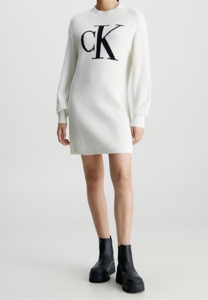 Nainen pukeutuneena valkoiseen ribbineulottuun neulemekkoon, jossa on suuri musta "cK"-logo, ja mustiin paksupohjaisiin nilkkureihin, seisoo valkoisella taustalla.