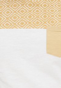 Επάνω τμήμα ένδυσης με κίτρινο και λευκό γεωμετρικό διαμαντένιο σχέδιο, μονόχρωμο λευκό ύφασμα από κάτω και μια μικρή μονόχρωμη κίτρινη τσέπη δεξιά.