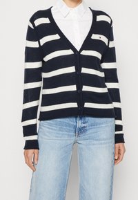 Cardigan blu navy con strisce orizzontali bianche, scollo a V, chiusura con bottoni e polsini a coste, abbinato a jeans azzurri.