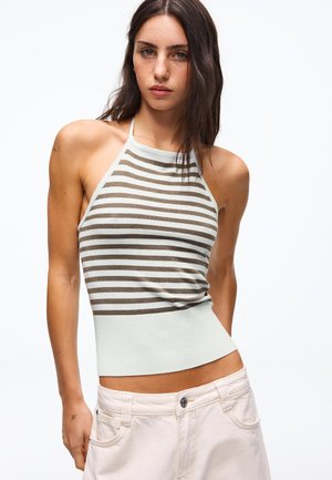 Nő hosszú sötét hajjal, ujjatlan pántos felsőt visel, amely vízszintes bézs és fehér csíkos, valamint világos színű nadrágot.