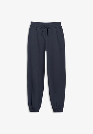 Jonge jongen staat met handen in de zakken, draagt een olijfgroene half-rits trui, marineblauwe joggingbroek, witte sokken en witte sneakers tegen een grijze achtergrond.