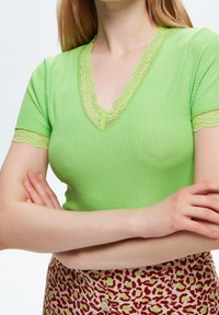 Donna con le braccia incrociate, che indossa un top verde a costine con scollatura a V e pizzo, e una gonna a fantasia con macchie rosse e verdi e bottoni bianchi.