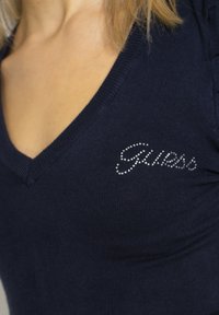 Femme portant un t-shirt bleu marine à col en V avec de petits strass formant le mot « Guess » sur le côté gauche de la poitrine, montrant une partie du cou et des cheveux.