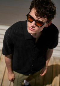 Black short-sleeve shirt with a textured surface, buttons down the front, paired with oversized brown-tinted sunglasses. Wood floor background.