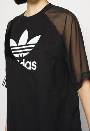 Osoba ubrana w czarną koszulkę Adidas z białym logo Trefoil, z przezroczystymi siateczkowymi rękawami i asymetrycznym wzorem warstwowej tkaniny.