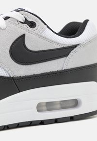 Grå mockasinsneaker i läder och svart med vitaAccenter, som har en synlig luftkudde, svart Nike-logotyp och texturerade meshsektioner.