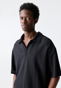 Polo shirt nera lavorata a maglia con un motivo testurizzato, vestibilità oversize, maniche corte e colletto classico con un'apertura a V sottile.