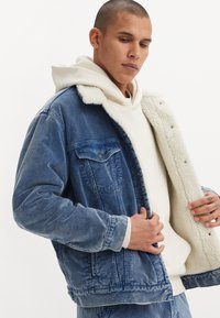 Denim jacket in light blue with a textured corduroy design. Features a white faux shearling collar and interior lining, and snap buttons.