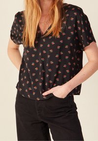 Donna che indossa una blusa nera a maniche corte con piccoli motivi floreali rossi e pantaloni neri, con una mano in tasca, adornata da collane d'oro a strati.