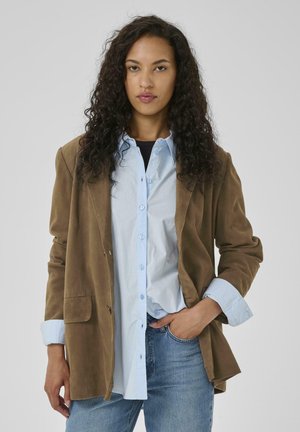 Brown suede blazer with a relaxed fit, featuring two front pockets, layered over a light blue button-up shirt. Model wears denim jeans.