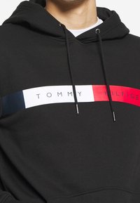 Felpa nera con cappuccio e una grande striscia orizzontale bianca, blu e rossa con il testo "TOMMY HILFIGER" in un colore a contrasto. Sono presenti dei cordini.