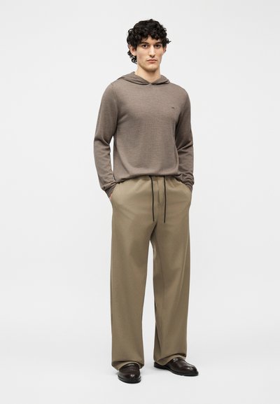 Νεαρός άνδρας με σγουρά μαλλιά, που φορά καφέ πουλόβερ με κουκούλα, μπεζ παντελόνι με κορδόνι και σκούρα καφέ παπούτσια σε απλό φόντο.