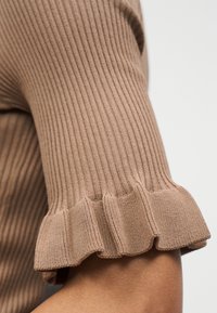 Manche d'un pull beige en maille côtelée avec un bord à volants, montrée en gros plan sur le bras d'une personne sur un fond uni.