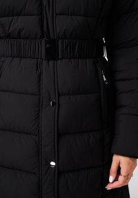 Zwarte gewatteerde jas met een taille accentuering door een glanzende gesp, drukknoopsluiting en zijzakken. De stof is gestructureerd en warm.