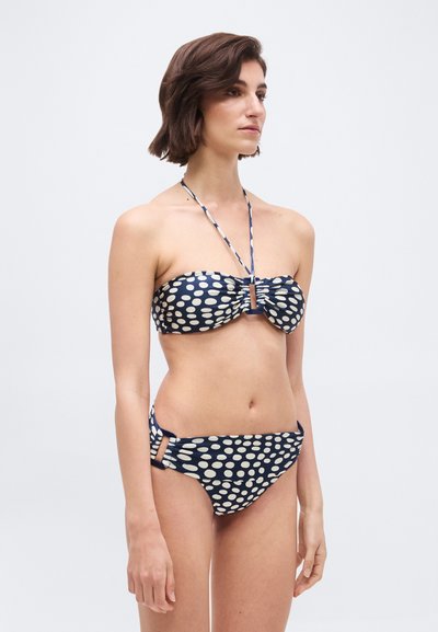 Γυναίκα που στέκεται με μπικίνι σε ναυτικό μπλε χρώμα με λευκές βούλες, τοπ χωρίς τιράντες με δέσιμο στον λαιμό και αντίστοιχα εσώρουχα μπικίνι με ανοίγματα στα πλαϊνά.