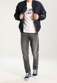 Sotilasaappaan takki vaalean graafisen t-paidan päällä, yhdistettynä harmaisiin farkkuihin ja sinivalkoisiin lenkkareihin. Raskas kangas ja rento malli.