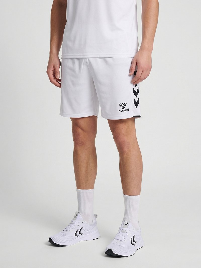 Mann trägt weiße Sportshorts, weiße Sportsocken und weiße Sneakers mit schwarzen Chevron-Designs, steht vor einem einfarbigen hellen Hintergrund.