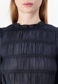 Navy blue, textured blouse with gathered fabric and pleats. Features a ruffled neckline and subtle sheen, suitable for various occasions.