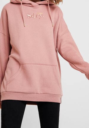 Vrouw draagt een oversized stofroze hoodie met reliëflogo en kangoeroezak, gecombineerd met zwarte broek tegen een lichte achtergrond.