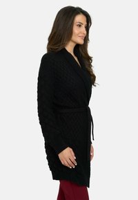 Cárdigan negro de punto con un patrón texturizado en las mangas, de frente abierto y con un cinturón atado a la cintura. Manga larga y longitud a medio muslo.