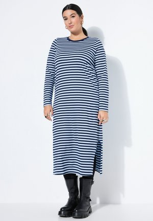 Kvinde iført en lang navy- og hvidstribet kjole med sideslid samt sorte leggings og sorte chunky støvler, der står mod en hvid baggrund.