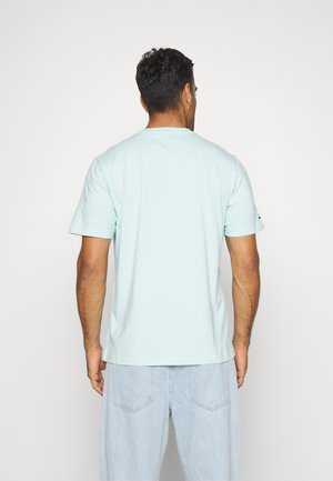 Mann mit kurzen dunklen Haaren, der ein hellblaues T-Shirt und hell gewaschene Jeans trägt, steht mit dem Rücken zur Kamera.