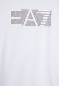 Camiseta blanca de algodón con un diseño de logo geométrico en blanco y negro que incluye "EA" y el texto "EMPORIO ARMANI" en el centro, con letras en relieve.