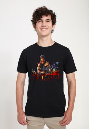 Ung mand med krøllet hår iført en sort T-shirt med en muskuløs karakter, der holder et stort våben, og ordet "RAMBO" i røde bogstaver.