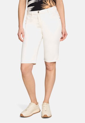Femme portant un short blanc jusqu'aux genoux, un haut à motif tropical et des baskets beige, debout les mains détendues le long du corps.