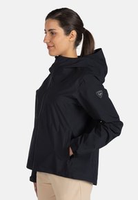Zwarte waterdichte jas met een capuchon, voorzien van een centrale rits, zijzakken en een logopatch op de linker mouw. Soepele textuur.