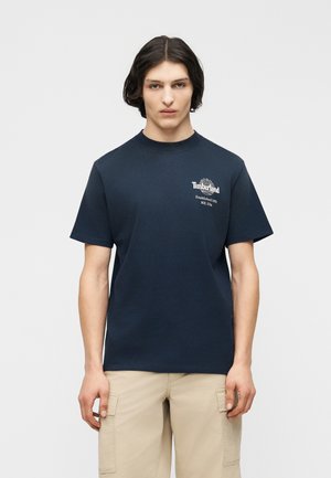 Junger Mann mit mittellangen dunklen Haaren trägt ein marineblaues Timberland-T-Shirt und beige Cargo-Shorts, steht vor einem schlichten hellen Hintergrund.
