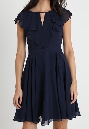 Femme portant une robe bleu marine longueur genou avec manches volantées, une profonde fente en V, une taille ajustée et une jupe plissée fluide.