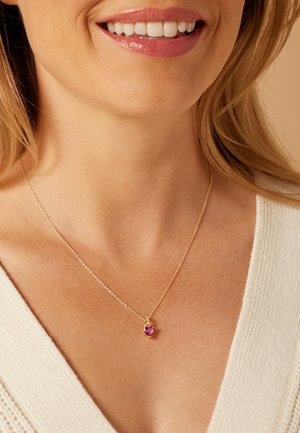 Goldene Kettenhalskette mit einem kleinen, herzförmigen rosa Edelsteinanhänger, präsentiert vor einem hellen Oberteil.