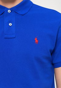 Plava polo majica izrađena od teksturiranog materijala, s kragnom, dva dugmeta i malim crvenim izvezenim logotipom na lijevoj strani prsnog koša.