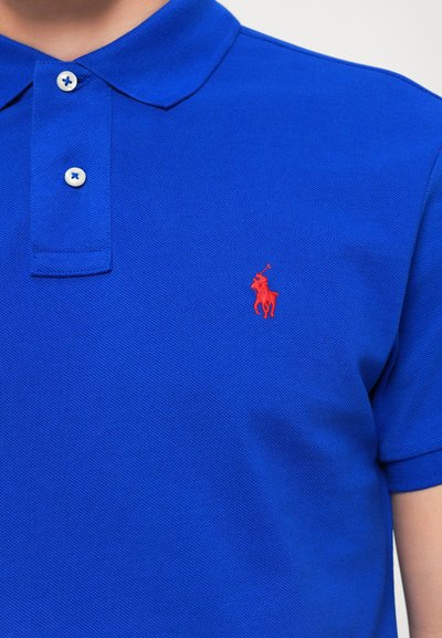 Zils polo krekls, veidots no strukturētas auduma, ar apkakli, divām pogām un nelielu sarkanu izšūtu logotipu kreisajā krūtīs.