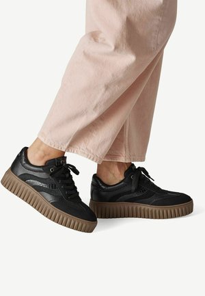 Persoon draagt lichtroze broek en zwarte sneakers met dikke bruine geribbelde zolen, gezien van opzij ter hoogte van de enkel.