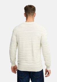 Pullover di colore crema chiaro in tessuto strutturato con un motivo ondulato, maniche lunghe e polsini e orlo a costine. Indossato con jeans in denim scuro.