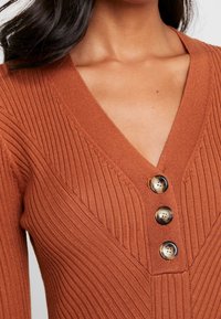 Hnědočervený pletený cardigan s V-výstřihem, vertikálním žebrovaným vzorem a třemi kulatými hnědými knoflíky vpředu.