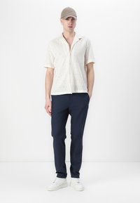 Kortermet bluse i kremfarge med blonder og knappelukking, kombinert med marineblå bukser og hvite joggesko. En beige caps fullfører antrekket.