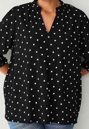 Blouse noire à pois blancs, manches trois-quarts à volants et petit col en V, portée avec un jean bleu.