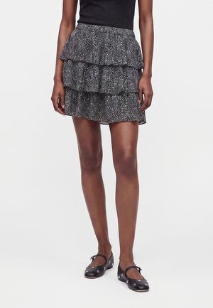 Femme portant une mini-jupe noire à volants avec des taches blanches, un haut noir et des chaussures plates noires avec des œillets métalliques.